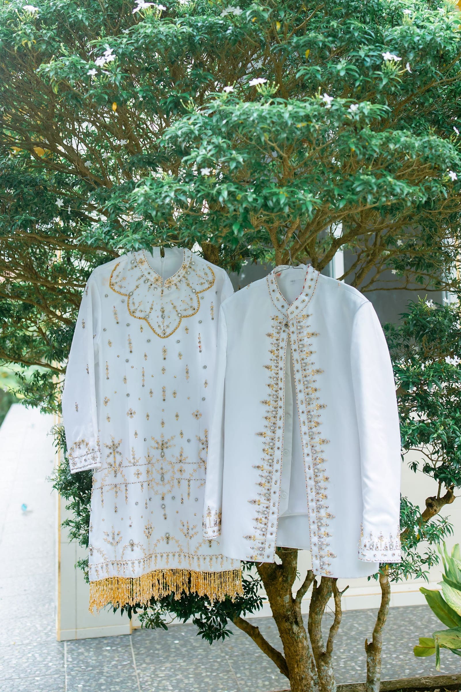 Two white kurtas with gold thread embroidery hanging from a tree in daylight